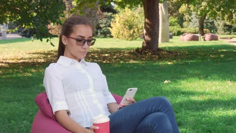 Young-woman-sitting-in-city-park-drink-coffee-and-browsing-on-phone