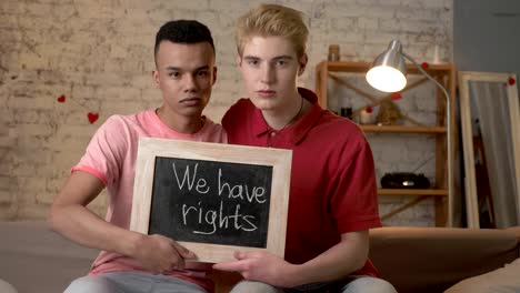 A-sad-international-gay-couple-is-sitting-on-the-couch-and-holding-a-sign.-We-have-rights.-Look-at-the-camera.-Home-comfort-on-the-background.-60-fps