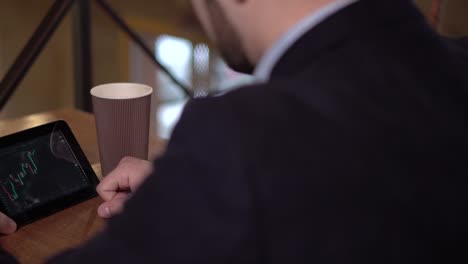 Young-businessman-monitors-trading-cryptocurrency-on-tablet-and-drinking-coffee-in-a-cafe