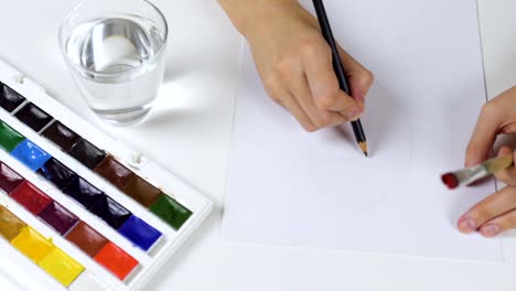 Unrecognizable-woman-draws-a-pencil-sketch-on-paper-Easter-card.-On-the-table-is-a-palette-with-paints-and-a-glass-of-water.