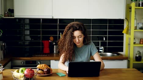 bastante-rizada-caucásica-joven-sentado-solo-a-la-mesa-en-idoors-amplias-cocina-trabajando-en-laptop-escuchando-música,-impresión-en-portátil-y-bailando