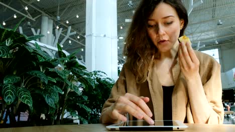 Young-businesswoman-uses-a-tablet-at-a-table-in-a-mall,-a-girl-eats-an-apple-and-works-at-a-cafe-remotely