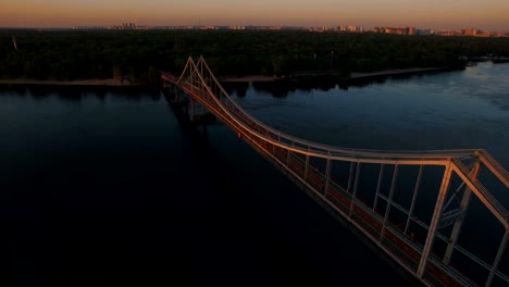 Pedestrian-bridge-over-the-river-near-the-city-at-sunset-aerial-drone-shoot