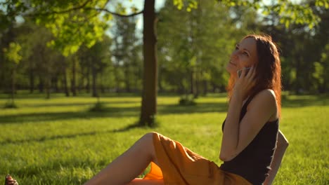 Happy-handsome-female-sitting-on-green-grass-and-have-conversation-via-digital-smartphone-device