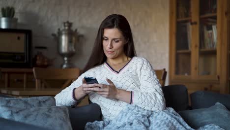 Frau-sitzt-auf-der-Couch,-eingewickelt-in-eine-Decke-und-SMS-auf-dem-Handy