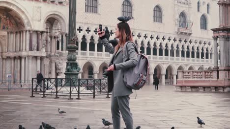 Happy-smiling-female-tourist-with-pigeons-sitting-on-her-arm-and-head-takes-selfie-on-city-square-in-Venice-slow-motion.