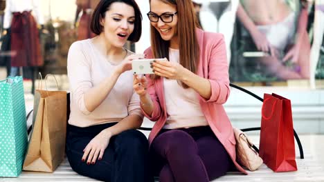 Emotional-girls-are-looking-at-smartphone-screen-chatting-and-laughing-sitting-on-bench-in-shopping-mall-with-bags-and-having-fun.-Modern-technology-and-communication-concept.