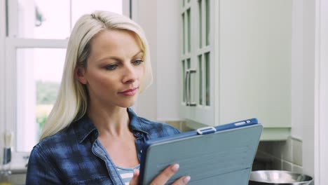 Beautiful-Blond-Woman-Scrolling-Through-Digital-Tablet-In-Kitchen