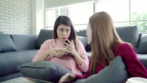 Asian-women-playing-pillow-fight-and-eating-popcorn-in-living-room-at-home,-group-of-roommate-friend-enjoy-funny-moment-while-lying-on-the-sofa.-Lifestyle-women-relax-at-home-concept.