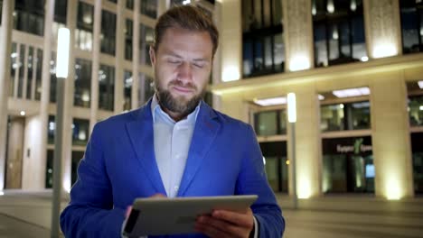 Carro-medio-tiro-de-guapo-empresario-adulto-mediano-navegando-por-Internet-en-tablet-PC-mientras-camina-al-aire-libre-en-la-calle-de-la-ciudad-de-noche