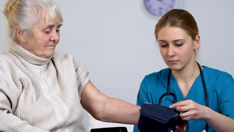 Young-nurse-measuring-disabled-elderly-female-blood-pressure-and-taking-records