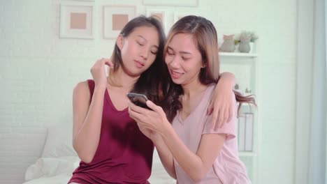 Pareja-feliz-lesbianas-jóvenes-mujeres-asiáticas-con-smartphone-control-de-medios-sociales-en-el-dormitorio-en-el-hogar,-disfrutar-de-momento-de-amor-mientras-está-acostado-en-la-cama-cuando-relajados.-Estilo-de-vida-LGBT-pareja-juntos-en-el-interior-co