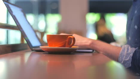 Hands-of-man,-male-digital-nomad-writing,-typing-on-laptop,-working-online-and-drinking-coffee-from-an-orange-cup-in-a-coffee-shop,-depth-shot