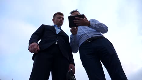 Low-angle-view-of-young-businessman-showing-a-presentation-on-screen-of-tablet-pc-to-his-colleague-in-city.-Successful-entrepreneurs-working-on-digital-tablet.-Blue-sky-at-background.-Slow-motion