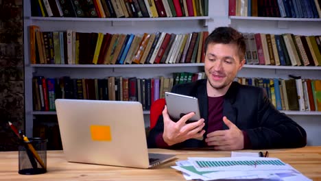 Attractive-adult-happy-businessman-having-a-video-call-on-a-tablet-sitting-in-front-of-the-laptop-in-library-indoors