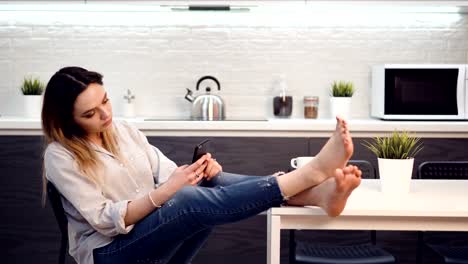 Young-girl-is-sitting-in-spacious-bright-kitchen-using-smartphone.