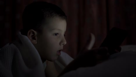 A-happy-boy-lies-in-bed-under-a-blanket-and-plays-on-a-tablet-in-a-game-in-the-dark.-The-face-of-the-child-is-lit-by-a-bright-monitor