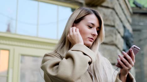 Portrait-of-cute-girl-in-headphones,-listening-to-music-and-browsing-on-mobile-phone-outddor.-Blogger-scrolls-through-social-media-on-device,-changing-tracks,-reading-news-on-app