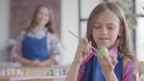 Portrait-of-cute-concentrated-girl-with-long-hair-in-blue-apron-painting-Easter-egg