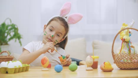 Nice,-cute-girl-is-having-fun-painting-an-Easter-egg.-Adorable-girl-is-colorizing-and-Ester-egg-.-Girl-with-beauty-spots-at-her-face-and-is-smiling-gently-with-cat's-whisker,-sitting-at-the-wooden-table-with-Easter-decorations.