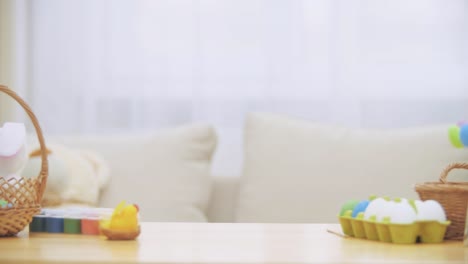 Little-playing-girl-with-bunny-ears-on-his-head-is-hiding-under-the-wooden-table,-full-of-Easter-decorations.-Girl-is-playing-with-an-Easter-white-bunny-on-the-table,-slolwly-showing-her-charming-face.