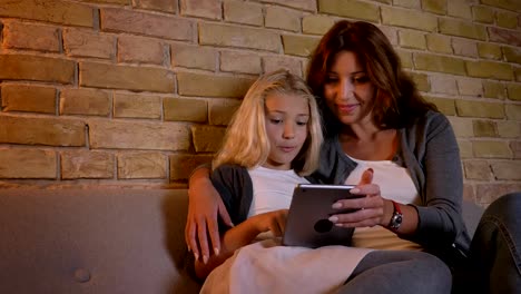 Closeup-shoot-of-young-pretty-caucasian-mother-and-her-small-blonde-daughter-using-the-tablet-together-and-browsing-the-web-sitting-on-the-couch