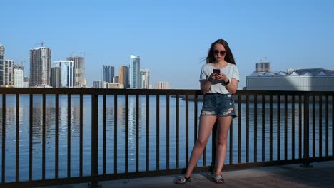 Beautiful-girl-with-long-hair-in-sunglasses-using-smartphone-app-at-sunset-river-quay-near-4K.