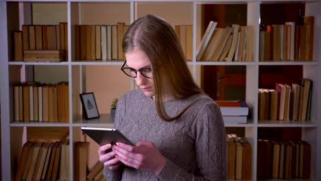 Nahaufnahme-Porträt-von-jungen-attraktiven-Studentin-in-Brille-mit-dem-Tablet-und-lächelnd-Blick-auf-die-Kamera-in-der-College-Bibliothek-drinnen