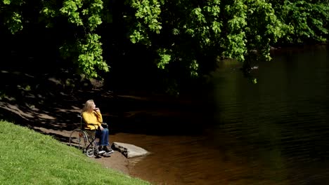 Mujer-mayor-en-silla-de-ruedas-mirando-a-la-vista-por-el-lago-en-el-parque-verde-en-el-día-de-verano-ventoso.-Inhabilitada-mujer-madura-contemplando-en-el-lago