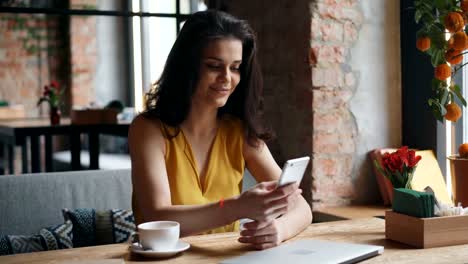 Charming-young-lady-holding-smartphone-touching-screen-smiling-in-modern-cafe