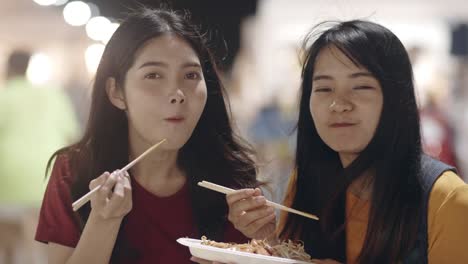 Asian-young-women-friends-travel-in-Bangkok,-Thailand,-beautiful-female-feeling-happy-walking-and-eating-Pad-Thai-at-The-Khao-San-Road.-Women-travel-eat-street-food-concept.-Slow-motion-shot.