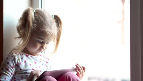 Blonde-kleine-Mädchen-mit-Tablet-PC.-Sitzen-auf-der-Fensterbank.-Kind-lächelnd-und-mit-Blick-auf-die-Kamera.-Kind-spielt-am-PC