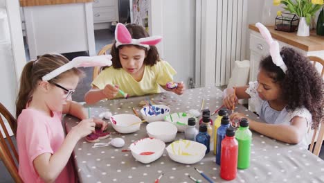 Grupo-de-chicas-sentadas-alrededor-de-la-mesa-de-la-cocina-con-orejas-de-conejo-decorando-huevos-para-Semana-Santa---tiro-en-cámara-lenta