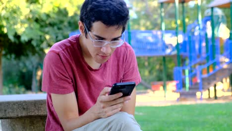 Joven-usando-su-teléfono-inteligente-sentado-en-un-banco-del-parque