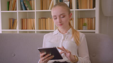 Retrato-de-primer-plano-de-joven-bonita-estudiante-rubia-caucásica-usando-la-tableta-mirando-la-cámara-sonriendo-felizmente-sentado-en-el-sofá-interior-en-el-apartamento