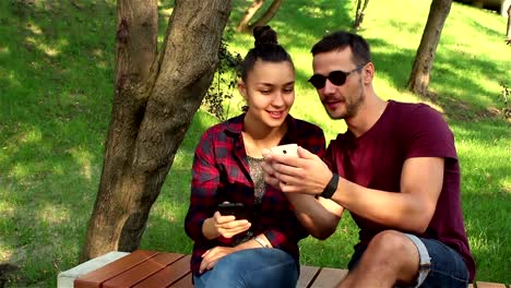 Couple-in-love-show-photos-on-their-smartphones-with-each-other,-sitting-on-a-park-bench.