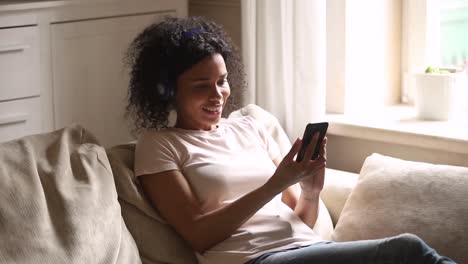 Feliz-joven-africana-usando-auriculares-inalámbricos-usando-teléfono-inteligente