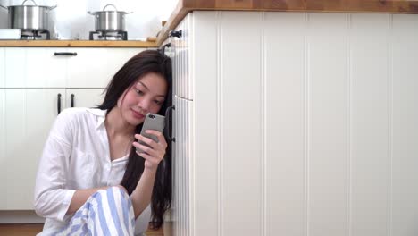 4K-Medium-dolly-shot-of-young-asian-woman-sitting-on-kitchen-floor-at-home-using-smartphone-with-wireless-technology-for-social-media-communication.-Asian-teenage-girl-enjoying-on-kitchen-floor-with-smartphone.