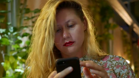 Pensive-girl-uses-a-smartphone,-writes-a-message,-chatting.-Woman-looking-for-information-in-the-phone-and-sad.-Sitting-in-restaurant-alone.-Evening-time,-lowlight.-Technology-and-entertainment-concept.-4k