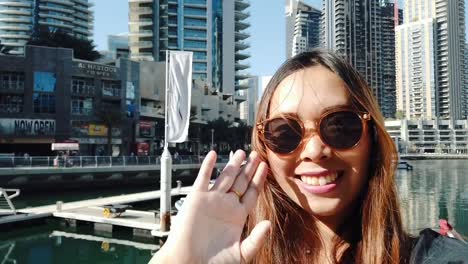 Cheerful-young-woman-takes-selfie-portrait-using-smartphone-sharing-travel-friends-showing-at-Dubai-marinaUnited-Arab-Emirates,-Dubai.