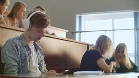 A-male-student-in-a-University-auditorium-listens-to-music-in-white-headphones-during-a-break-without-paying-attention-to-others.-A-lot-of-people-in-the-audience-are-talking