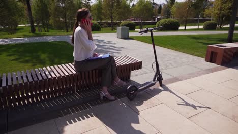 Una-mujer-de-negocios-hablando-por-teléfono,-sentada-en-un-parque-al-lado-de-una-moto.-Joven-mujer-moderna-con-atuendo-de-negocios.-Conversación-al-aire-libre