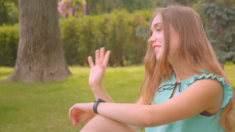 Closeup-portrait-of-young-cute-long-haired-beautiful-female-sitting-on-green-grass-using-smart-watch-in-park-outdoors