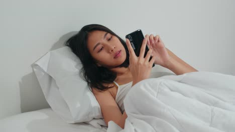 Junge-asiatische-Frau-mit-Smartphone-Check-Social-Media-Gefühl-glücklich-lächeln,-während-auf-dem-Bett-liegen-duponieren,-nachdem-sie-am-Morgen-aufwachen,-Schöne-attraktive-hispanische-Mädchen-lächelnd-entspannen-im-Schlafzimmer-zu-Hause.