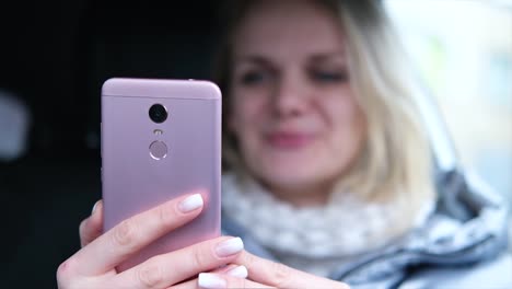 Verschwommene-blonde-Frau-Bloggerin-in-Winterkleidung-sitzen-im-Auto-und-im-Gespräch-mit-Anhängern,-Live-Streaming,-Blick-auf-Smartphone-Bildschirm.-Fokus-auf-Telefon