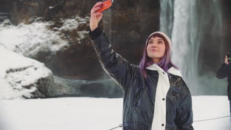 schöne-junge-Frau-in-der-Nähe-eines-Wasserfalls-in-Island,-macht-Fotos-am-Telefon,-freut-sich-und-macht-ein-Selfie