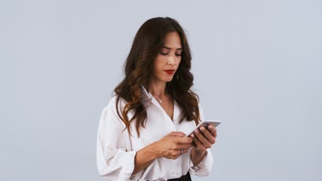 Brunette-woman-in-white-shirt-is-typing-on-smartphone,-smiling,-rejoicing-and-crossing-her-fingers-while-posing-on-gray-background.-Close-up