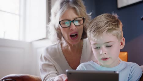 Close-Up-Of-Grandson-With-Grandmother-Sitting-In-Chair-Playing-On-Digital-Tablet-At-Home-Together