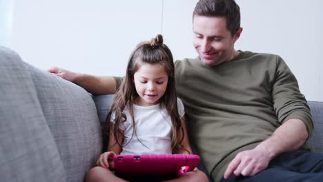 Vater-und-Tochter-sitzen-auf-Sofa-zu-Hause-spielen-zusammen-auf-digitalen-Tablet-in-rosa-Fall-zu-Hause