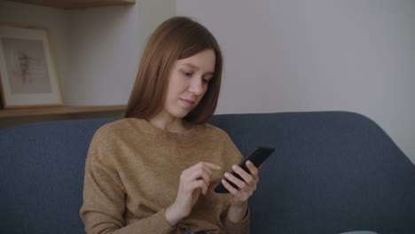 Young-woman-sitting-at-desk-at-home-office-distracted-from-work-holding-smart-phone-using-social-networking-website-chatting-with-friend-remotely-using-modern-device-and-internet-connection